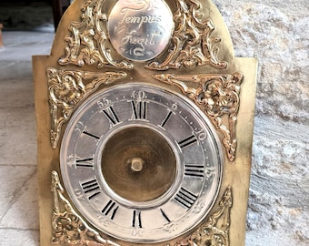 May include: A brass clock face with Roman numerals and a silvered center. The clock face is surrounded by an ornate brass frame with floral designs. The top of the frame features a circular plaque with the inscription "Tempus Fugit".
