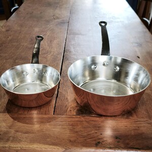 May include: Two copper-bottomed saucepans with riveted construction and black handles. The pans have a shiny, silver interior and a warm, reddish-brown exterior. One pan is smaller than the other, and both are sitting on a wooden surface.