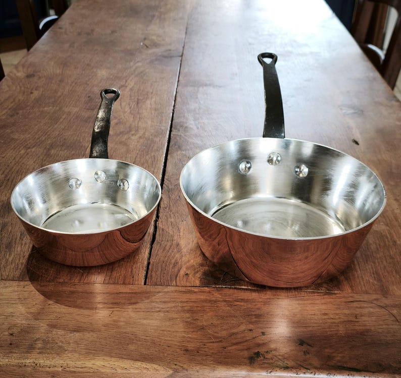 May include: Two copper-bottomed saucepans with riveted construction and black handles. The pans have a shiny, silver interior and a warm, reddish-brown exterior. One pan is smaller than the other, and both are sitting on a wooden surface.