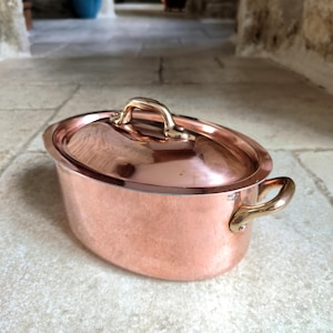 May include: Oval copper cooking pot with lid and brass handles. The pot has a shiny, reflective surface and is on a stone surface. The lid has a brass handle. A classic kitchenware item.