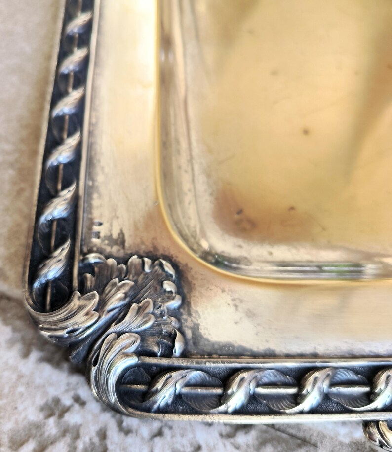 May include: Close-up of a silver serving tray with an ornate, detailed border. The tray has a rectangular shape and a smooth, polished surface.