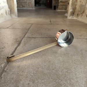 May include: A copper and brass ladle with a long handle and a rounded bowl. The bowl is silver-colored on the inside and copper on the outside. The handle is brass-colored and has a hook at the end.