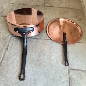 May include: A set of two copper pots with black handles. The larger pot has a lid. The pots are shiny and have a traditional design.