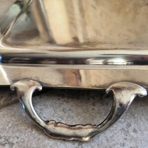 May include: Close-up of a silver serving tray with ornate handles. The tray is made of polished metal and has a reflective surface.