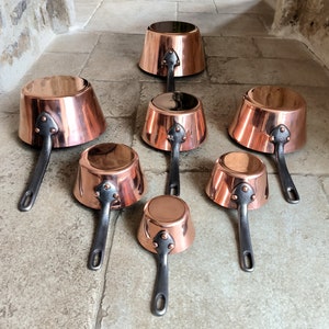May include: A set of seven copper pots with black handles. The pots are arranged in a pyramid shape, with the largest pot at the top and the smallest pot at the bottom.
