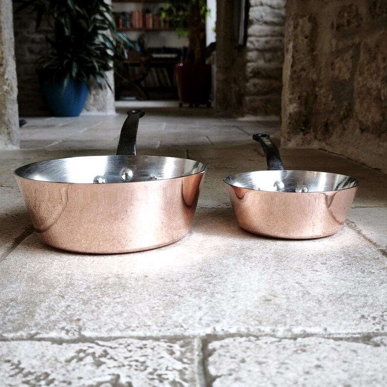 May include: Two copper saucepans with black handles. The pans have a shiny copper exterior and a silver interior. The pans are sitting on a stone surface. The larger pan is approximately 20 cm in diameter.