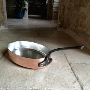 Pode incluir: Frigideira oval de cobre com interior prateado e cabo preto. O cabo é fixado à frigideira com rebites. A frigideira está sobre uma superfície de pedra. Medidas em centímetros.
