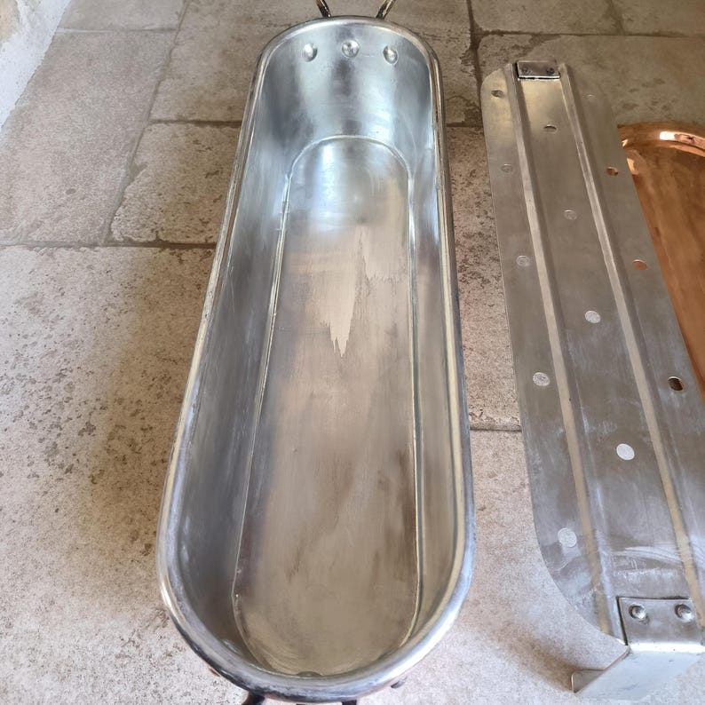 Puede incluir: Una ba&ntilde;era vintage de metal cepillado, de forma ovalada y alargada. Tiene dos asas y est&aacute; acompa&ntilde;ada de un panel met&aacute;lico desmontado. El dise&ntilde;o general sugiere un enfoque en la funcionalidad y la durabilidad. La ba&ntilde;era mide unos 150 cm de largo.