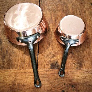 May include: Two copper saucepans with dark metal handles on a wooden surface. The pans have a shiny, reflective surface and a classic design. The handles are riveted to the copper bodies.