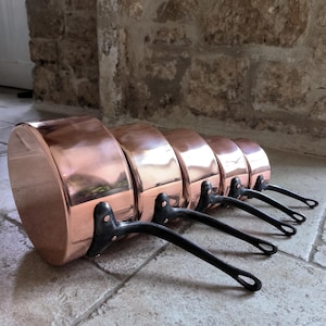 Peut inclure: Ensemble de six casseroles en cuivre avec des poignées noires. Les casseroles sont disposées en rangée, la plus grande casserole à l'avant et la plus petite casserole à l'arrière.