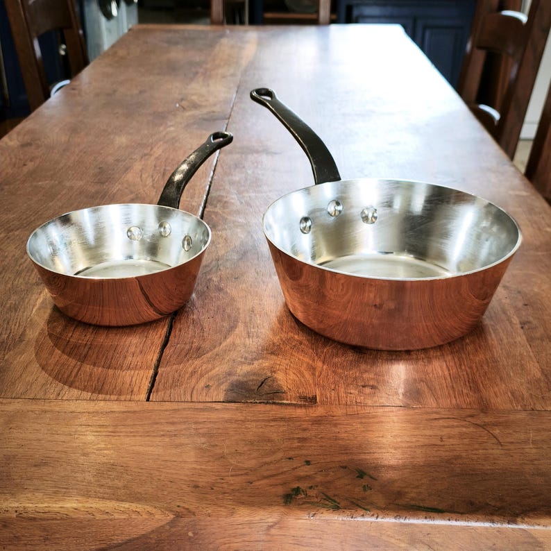 May include: Two copper saucepans with black handles on a wooden table. The pans have a shiny, silver interior and a copper exterior. The handles are curved and attached with rivets. The pans are different sizes.