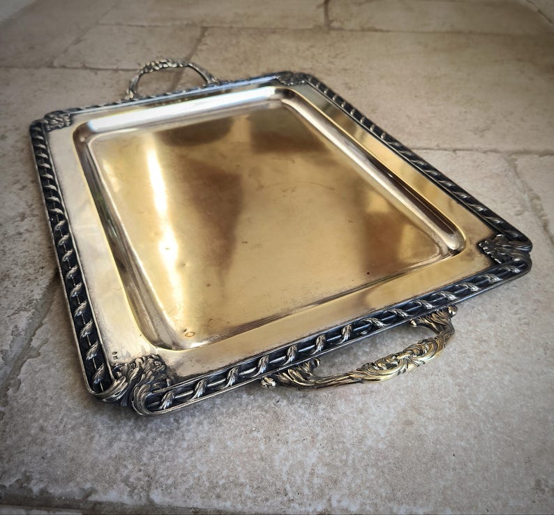 May include: A silver serving tray with a black rope design around the edge and two ornate brass handles.