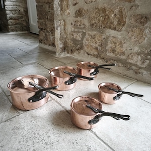 May include: Five copper saucepans with black handles, arranged on a stone floor. The shiny copper pots reflect the light. The background features a stone wall. The pans are of varying sizes.