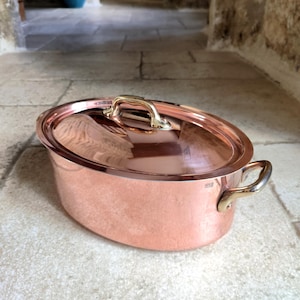 May include: A copper oval cooking pot with a matching lid. The pot has two side handles and a top handle, all in a gold tone. The pot is sitting on a stone surface.
