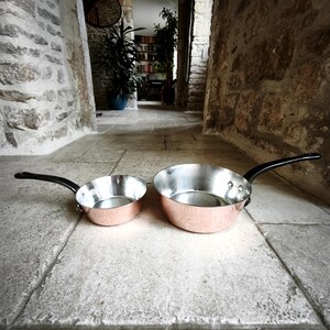 May include: Two copper pans with silver interiors and black handles. The pans are different sizes, with one smaller than the other. The pans are on a stone surface.