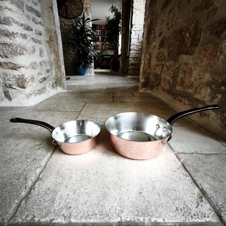 May include: Two copper pans with silver interiors and black handles. The pans are different sizes, with one smaller than the other. The pans are on a stone surface.