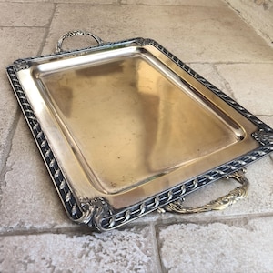 May include: A silver serving tray with a decorative border and two handles. The tray is made of metal and has a shiny finish.