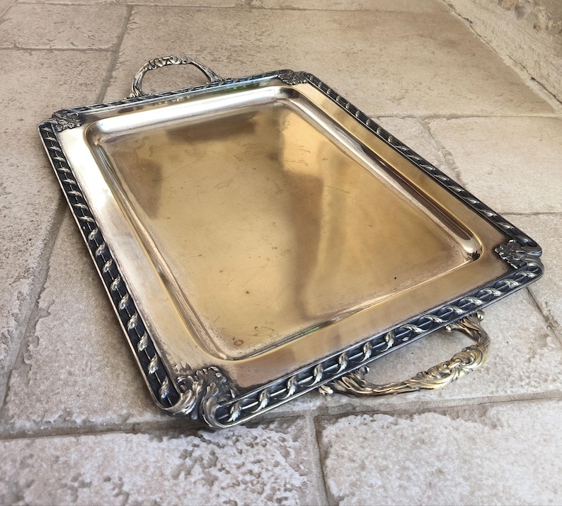 May include: A silver serving tray with a decorative border and two handles. The tray is made of metal and has a shiny finish.