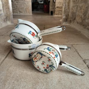 Peut inclure: Ensemble de casseroles en porcelaine blanche avec des motifs floraux et de volutes rouges, bleus et verts. Chaque casserole a un bord en métal et une poignée blanche avec un motif à pois verts. Les casseroles sont empilées.