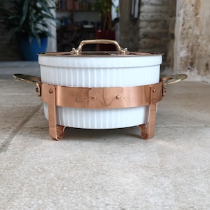 May include: A white, ribbed ceramic casserole dish with a copper-colored metal band and legs. The dish has a gold-colored handle on the lid and side handles. The casserole dish is designed for serving food.