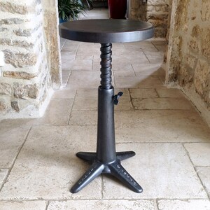 May include: A dark gray, adjustable height stool with a round seat. The stool features a threaded central column, a four-point base, and a lever for height adjustment. The design has an industrial, vintage aesthetic.