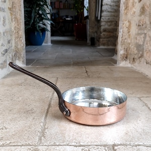 Op de afbeelding: Een koperen koekenpan met een lange, donkere handgreep. De binnenkant van de pan is zilverkleurig. De pan staat op een stenen ondergrond. De handgreep is met klinknagels bevestigd. Diameter ca. 20 cm.