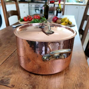 Pode incluir: Uma panela redonda de cobre martelado com tampa a condizer. A panela possui duas pegas laterais robustas de latão rebitado, e a tampa inclui uma pega central de latão. A superfície exterior apresenta uma textura distinta com marcas de martelo.