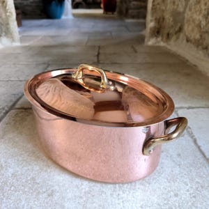 May include: A copper oval cooking pot with a matching lid. The pot has brass handles on the sides and the lid. The pot is sitting on a stone surface. The pot is a shiny, reflective copper color.