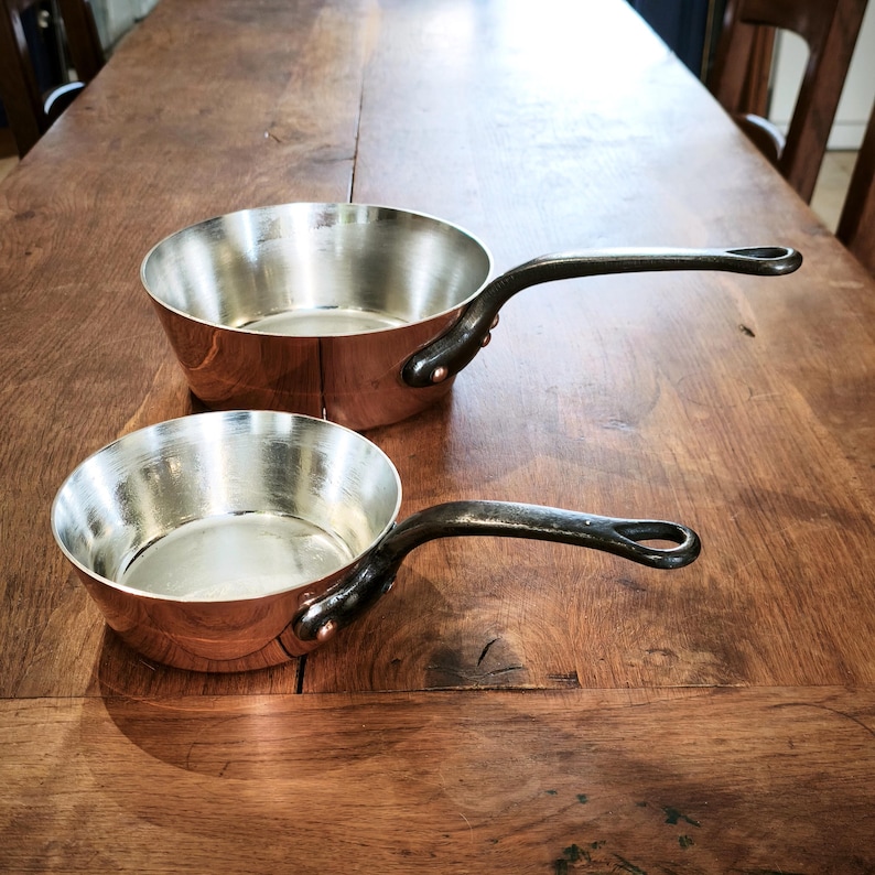 May include: Two copper saucepans with silver interiors and dark handles sit on a wooden surface. The pans have a classic design, with a rounded body and a long, curved handle. The copper exterior gives them a warm look. Ideal for cooking.