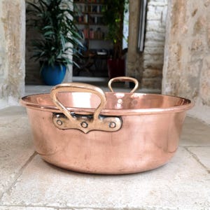 May include: A copper bowl with two brass handles. The bowl has a rounded shape and a polished finish, reflecting the surrounding light. The handles are attached with rivets. The bowl is sitting on a stone surface.