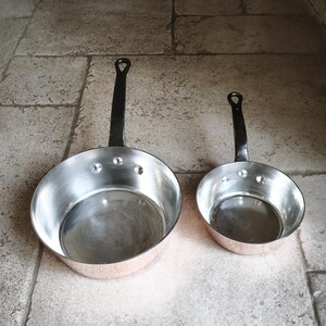 May include: Two copper-bottomed pans with riveted construction and black iron handles. The pans have a classic design, with a rounded shape and a flared rim. The handles are long and sturdy, with a loop at the end for hanging. The pans are of different sizes.