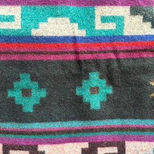 May include: Close-up of a woven textile with a geometric pattern. The design features repeating shapes in teal, white, purple, red, and black. The textile has a textured appearance, suggesting a wool or similar material. The overall aesthetic is reminiscent of Southwestern or Native American designs.