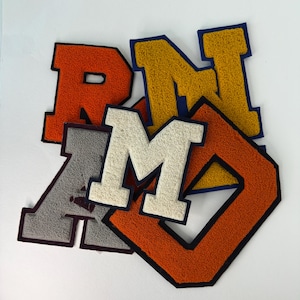 May include: A collection of five varsity-style letter patches featuring a textured chenille finish. The letters include an orange R, a yellow N, a grey A, a white M, and an orange D, each outlined with a dark blue or black border.