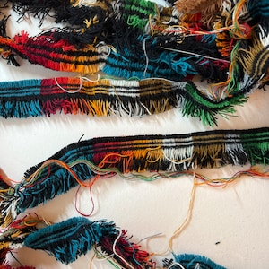 Puede incluir: Una colección de tiras de adorno de tela tejida multicolor con un borde deshilachado. El adorno muestra un patrón repetitivo de hilos negros, blancos, rojos, naranjas, amarillos y verde azulado, con hebras de hilo sueltas y enredadas.