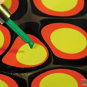 May include: A green dropper bottle with a green tip is being used to add a drop of green paint to a yellow circle surrounded by a red ring. The circle is on a black surface with other yellow circles with red rings.
