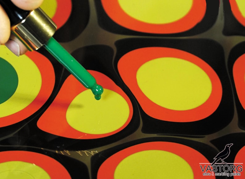 May include: A green dropper bottle with a green tip is being used to add a drop of green paint to a yellow circle surrounded by a red ring. The circle is on a black surface with other yellow circles with red rings.
