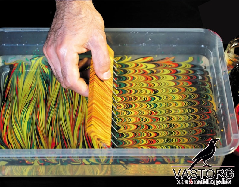 May include: A hand holding a wooden tool with metal pins over a tray of water with swirling yellow, green, red, and black paint. The paint is creating a marbled effect. The tray has the text "VASTORG ebru & marbling paints" on the bottom.