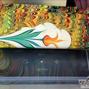 May include: A sheet of paper with a colorful marbled design is being submerged into a clear plastic container filled with water. The paper has a white center with a green and orange flower design. The container is surrounded by bottles of paint.