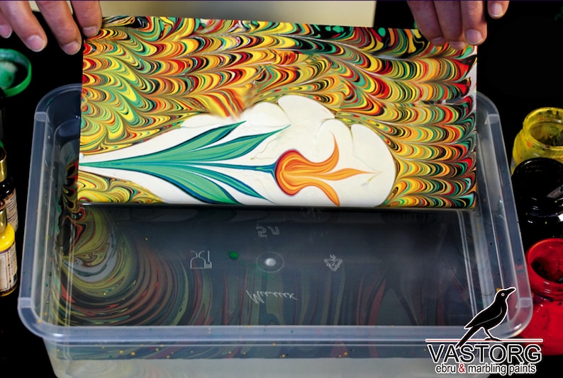 May include: A sheet of paper with a colorful marbled design is being submerged into a clear plastic container filled with water. The paper has a white center with a green and orange flower design. The container is surrounded by bottles of paint.