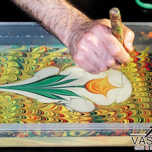 May include: A person uses a tool to create a swirling pattern in a tray of water with yellow, green, red, and black paint. The paint creates a flower-like design in the water. The text "VASTORG ebru & marbling paints" is visible in the bottom right corner of the image.
