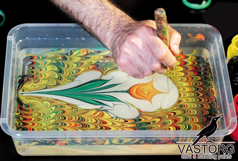 May include: A person uses a tool to create a swirling pattern in a tray of water with yellow, green, red, and black paint. The paint creates a flower-like design in the water. The text "VASTORG ebru & marbling paints" is visible in the bottom right corner of the image.