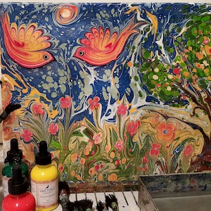 May include: A colorful abstract painting on canvas with two red and orange birds, a green tree, and flowers. The painting is on a table with bottles of paint and brushes.