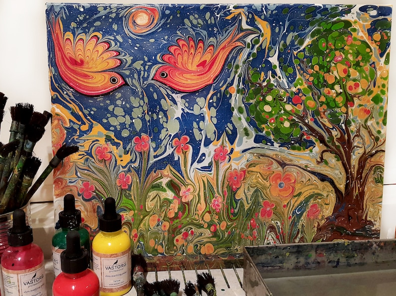 May include: A colorful abstract painting on canvas with two red and orange birds, a green tree, and flowers. The painting is on a table with bottles of paint and brushes.