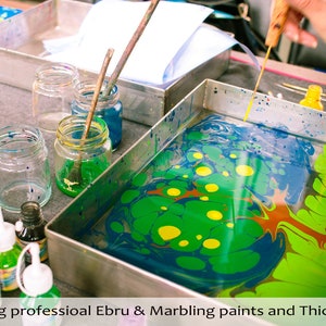 May include: A metal tray with green, blue, yellow, and red paint swirling in water. Jars of paint and a wooden tool are on the table next to the tray.  Vastorg professional Ebru & Marbling paints and Thickener.