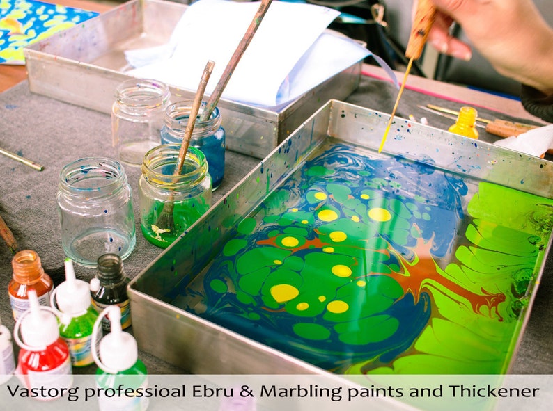 May include: A metal tray with green, blue, yellow, and red paint swirling in water. Jars of paint and a wooden tool are on the table next to the tray.  Vastorg professional Ebru & Marbling paints and Thickener.
