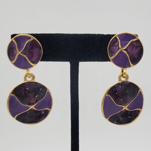 May include: A pair of gold tone dangle earrings with purple enamel. The earrings feature two round discs with a geometric pattern.