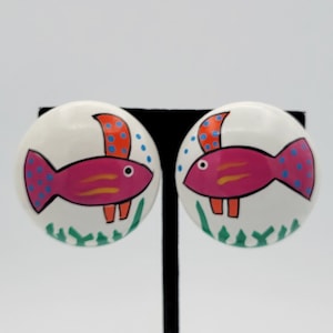 May include: A pair of white clip-on earrings with a pink fish design. The fish has orange fins, yellow stripes, and blue dots. The fish is surrounded by green seaweed.