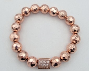 May include: Rose gold beaded bracelet featuring faceted, round beads. The bracelet has a rectangular clasp adorned with small, clear crystals. The beads have a hammered texture, giving them a unique, eye-catching appearance.