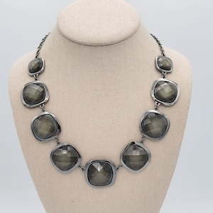 Può includere: Una collana a catena d'argento con sette grandi pietre grigie scure sfaccettate. Le pietre sono incastonate in castoni d'argento e sono disposte in uno schema sfalsato.