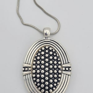 Puede incluir: Un collar de cadena de plata con un colgante ovalado. El colgante tiene una superficie texturizada con un patrón de pequeños círculos.